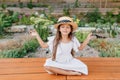 Pretty brunette female kid in straw hat sitting near flowerbed in lotus pose with eyes closed. Little girl in white Royalty Free Stock Photo