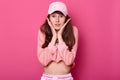 Pretty brunette female with wide open eyes, wears rosy shortened hoodie and cap, keeps palms on cheeks. Model stands on Royalty Free Stock Photo