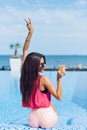 Pretty brunette girl with long hair is sitting near pool. She wears rose T-shirt, shorts, sunglasses. She holds drink in