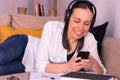 Young woman smiling with her smartphone studying stretched out on the sofa at home listening to music through headphones. Royalty Free Stock Photo