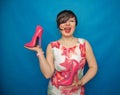 Pretty caucasian adult woman in white dress with flowers holding a pink Shoe with a very high heel on a blue solid Studio backgrou