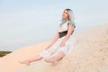 Young beautiful Caucasian woman in white dress and brown leather waistcoat posing in desert landscape with sand. Royalty Free Stock Photo