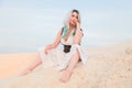 Young beautiful Caucasian woman in white dress and brown leather waistcoat posing in desert landscape with sand. Royalty Free Stock Photo