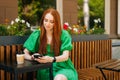 Pretty Caucasian young woman using browsing smartphone looking at screen, sitting at table with coffee cup in outdoor Royalty Free Stock Photo