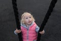 Pretty cheerful toddler girl with blond curly hair and big blue eyes happily swinging on a swing, made of black rope Royalty Free Stock Photo