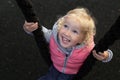 Pretty cheerful toddler girl with blond curly hair and big blue eyes happily swinging on a swing, made of black rope Royalty Free Stock Photo