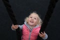 Pretty cheerful toddler girl with blond curly hair and big blue eyes happily swinging on a swing, made of black rope Royalty Free Stock Photo