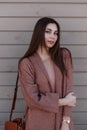 Pretty comely young woman with beautiful hair in long youth spring coat with leather fashionable handbag stands near wooden
