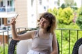 Pretty curly smiling young girl taking a selfie on smartphone on the apartment balcony. Video chatting with friends Royalty Free Stock Photo