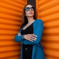 Pretty cute hipster young urban woman in trendy clothes in stylish sunglasses is posing outdoors near a bright orange metal wall. Royalty Free Stock Photo