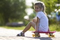 Pretty cute little blond girl in white shorts and T-shirt sitting on pink skateboard and smiling in camera on a light blurred sum Royalty Free Stock Photo