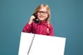 Pretty, cute little girl in pink shirt, black trousers and glasses holds empty poster and having a phone talk Royalty Free Stock Photo