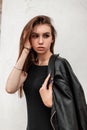 Pretty cute young woman in an autumn leather black jacket in a fashionable black dress straightens hair standing near a white wall Royalty Free Stock Photo