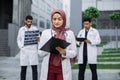Lady doctor in hijab writing notes on clipboard, standing ouside clinic with colleagues behind Royalty Free Stock Photo