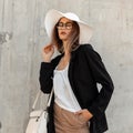 Pretty elegant in fashion glasses in beautiful sexy top in black blazer in summer hat with trendy handbag stands near vintage wall Royalty Free Stock Photo