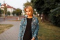 Pretty fashionable young woman blonde in blue vintage denim jacket with trendy leather handbag in dress posing on the street Royalty Free Stock Photo