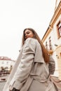 Pretty fashionable young woman in a stylish trench coat walks down the street and turns back. Modern trendy girl fashion model