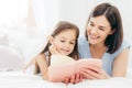 Pretty female mother and her small daughter read interesting book in bedroom, lie on comfortabled bed during morning. Lovely mum r Royalty Free Stock Photo