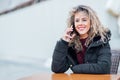 Pretty female speaking for smart phone sits in outdoor cafeteria