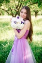 Pretty gentle young girl in a purple dress poses in an apple garden with a bouquet in her hands. Professional makeup and