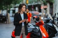 Pretty girl in business clothes holds smartphone background of rent mopeds in city of summer street outdoors, active tourist