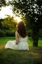 Back of a young woman with curly long hair sitting on the green grass in the park at summertime, admiring the sunset