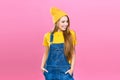 A pretty girl in denim overalls and a yellow hat on a pink background. Fashionista lady student. Bright trendy studio