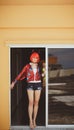Pretty girl in devil costume standing in a gloomy corridor by the window. Wears a red wig with horns, denim shorts and