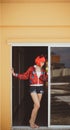 Pretty girl in devil costume standing in a gloomy corridor by the window. Wears a red wig with horns, denim shorts and