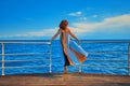 Pretty girl in flowing long dress stands on edge of pier.