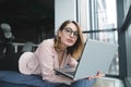 Pretty girl in the glasses lies at the window on the sofa with a laptop in her hands and looks at the camera