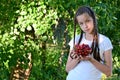 pretty girl with pigtails in white T-shirt is standing in garden and holding glass vase with ripe, juicy cherries in her hands. Royalty Free Stock Photo