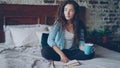 Pretty girl reading book sitting on bed at home and holding cup with drink enjoying free time and resting. Young people Royalty Free Stock Photo