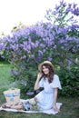 Pretty girl in a straw hat sits on a plaid and happily spends time on a picnic in the park with a lilac in the background. Concept