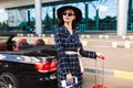 Pretty girl in sunglasses and black hat holding passport with flight ticket dreamily looking aside near airport with Royalty Free Stock Photo