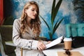 Pretty girl with wavy hair in striped trench coat sitting with book in hands and coffee to go on table while spending