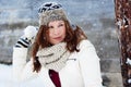 Pretty and fierce girl in winter clothing, scarf and mittens throws a snow ball in front of a wooden barn