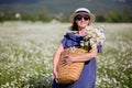 Beautiful woman with basket of flowers over chamomile field. Carefree happy brunette woman in straw hat in blue dress.