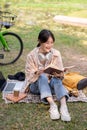 A pretty and happy young Asian woman is relaxing and reading a book in a green park on a bright day Royalty Free Stock Photo