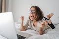 Pretty happy young curly woman with glasses spends time online shopping in bed, with a credit card in her hands with laptop, Royalty Free Stock Photo