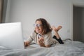 Pretty happy young curly woman with glasses spends time online shopping in bed, with a credit card in her hands with laptop, Royalty Free Stock Photo