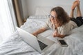 Pretty happy young curly woman with glasses spends time online shopping in bed, with a credit card in her hands with laptop, Royalty Free Stock Photo