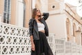 Pretty happy young woman model in elegant stylish clothes is standing and cute smiling near a vintage fence in the city on a warm Royalty Free Stock Photo