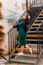 Pretty hipster girl is standing on iron stairs, Happy beautiful young woman, green dress, black jacket, urban background, street Royalty Free Stock Photo