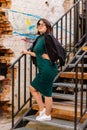 Pretty hipster girl is standing on iron stairs, Happy beautiful young woman green dress, black jacket, urban background, street Royalty Free Stock Photo