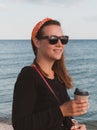 Pretty hipster woman in bandana long hair walking on sea background with coffee cup lifestyle real life travel photo