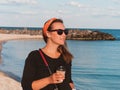 Pretty hipster woman in bandana long hair walking on sea background with coffee cup lifestyle real life travel photo