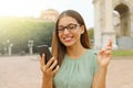 Pretty hopeful girl with glasses crossing fingers and holding a smart phone waiting for good news outdoor. Young woman with