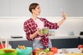 Pretty housewife taking selfie or making a video call using her smartphone while cooking fresh salad wearing a plaid Royalty Free Stock Photo