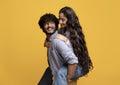 Pretty indian lady piggybacking her boyfriend, smiling guy carrying his girlfriend on his back, yellow background Royalty Free Stock Photo